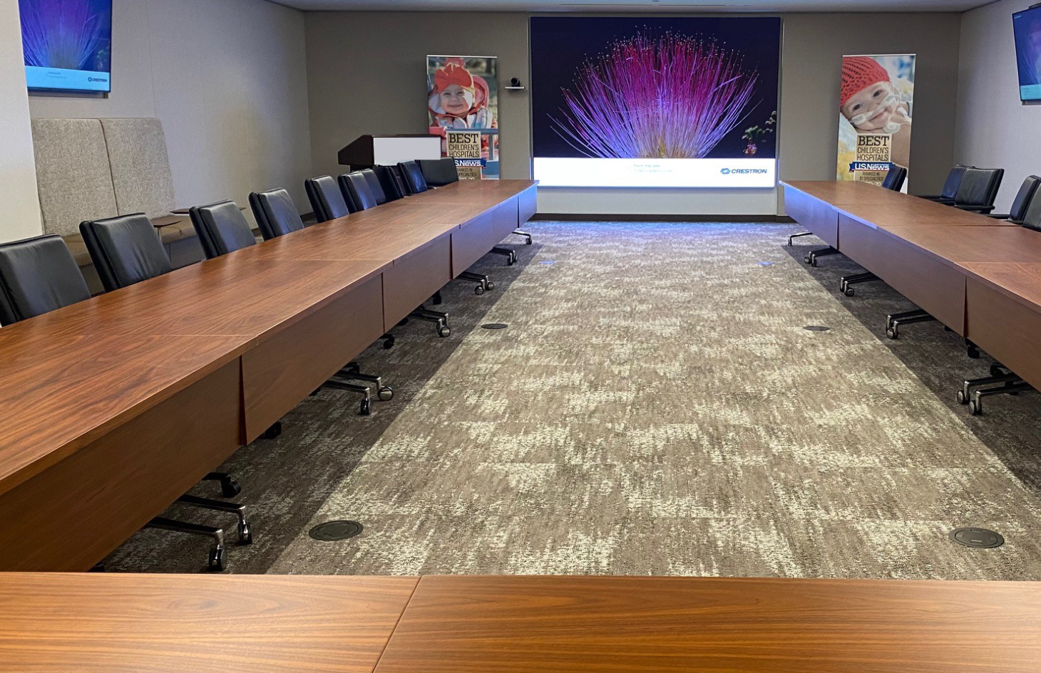 A long, rectangular conference room with a large presentation screen at the far end, flanked by posters featuring children’s images. Rows of wooden tables and black chairs face the screen.