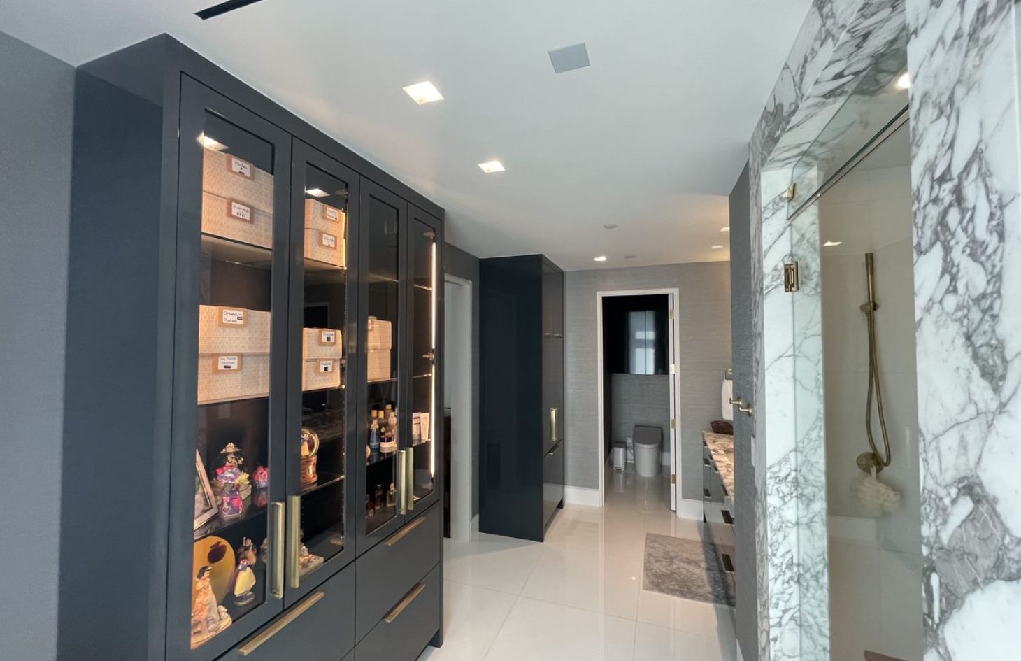 A modern, luxury bathroom featuring a glass-enclosed marble shower, sleek cabinetry with glass display doors, and a spacious vanity area. The space has recessed lighting, polished tile floors, and organized storage, creating a clean and upscale design.