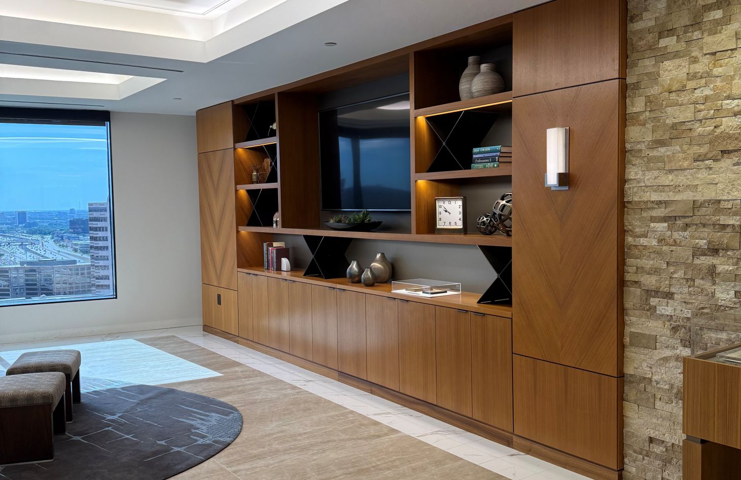 A modern living space with a custom-built-in wooden entertainment wall featuring shelves, decorative items, and a mounted television. The room includes a large window with a city view, soft lighting, and contemporary design elements such as stone accent walls and clean-lined furniture.