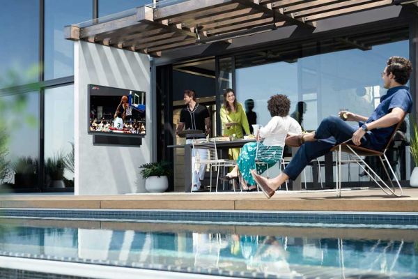 An outdoor patio area where several people watch a basketball game on a wall-mounted TV. The space includes modern seating, a pergola, and a pool in the foreground.